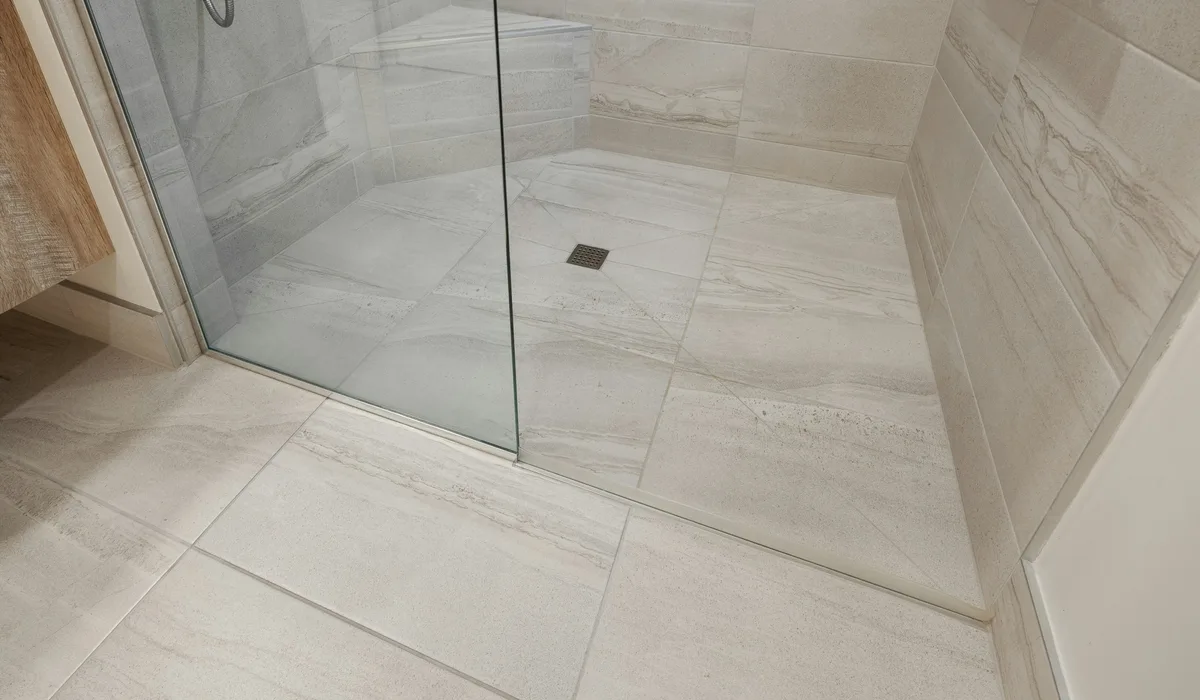 Curbless shower with light floor tile sloped toward a square drain behind a glass panel
